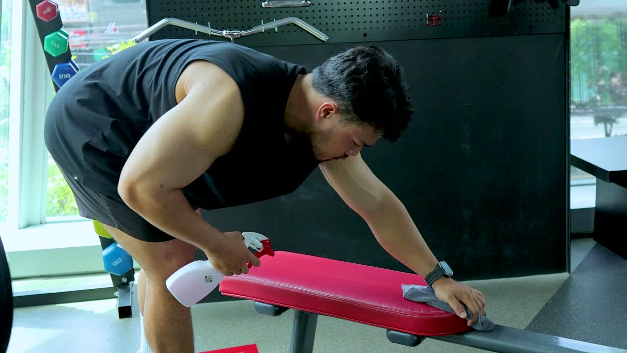 Athletic man cleans gym bench using disinfectant spray and cloth in bright, modern fitness center