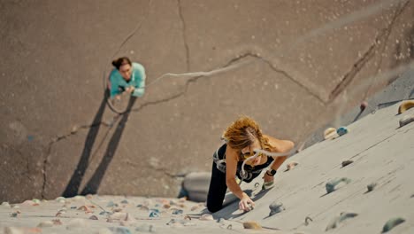 Top-view-of-a-blonde-girl-in-a-black-sports-summer-uniform-and-special-equipment-with-a-belay-climbing-up-a-white-climbing-wall-with-multi-colored-ledges.-And-below-on-the-ground-stands-her-friend-who-belays-the-girl-and-holds-the-rope-to-maintain-the-belay-during-the-climb-and-training-rock-climbin