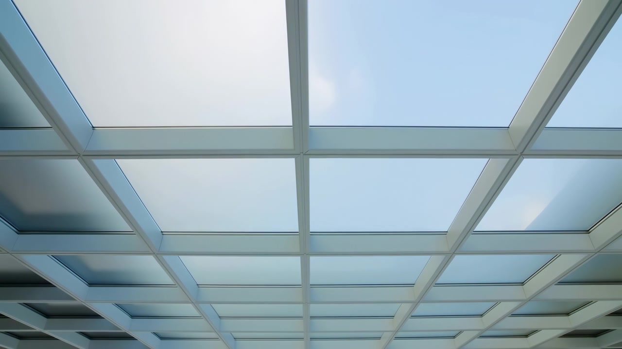 Upward view of modern glass skylight ceiling with white grid structure, large pale blue panes, clean architectural lines, smooth reflective surfaces, bright daylight, and airy minimalist atmosphere
