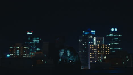 friends using smartphone mobile technology hanging out on rooftop at night enjoying weekend chatting sharing social media messages in urban city skyline background