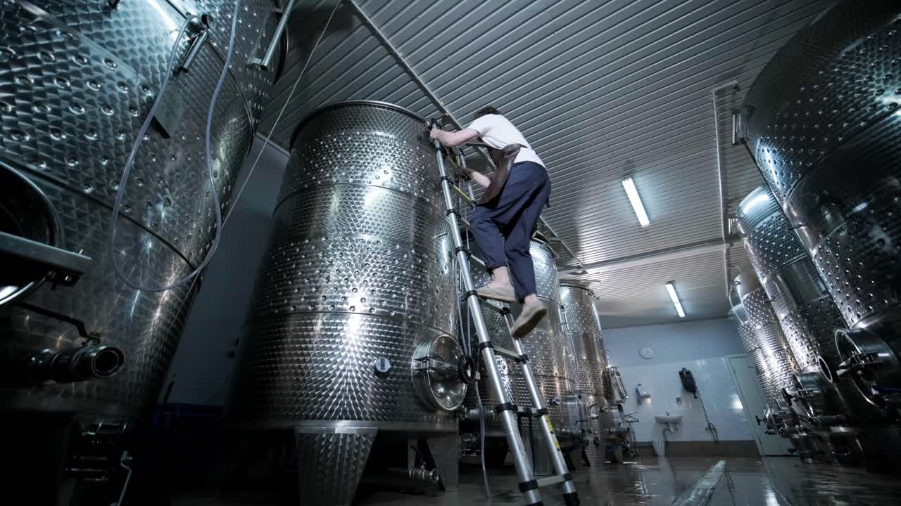 A happy successful farmer or winemaker is walking in the middle of wine tanks and checking a fermentation grade of high quality wine production in a modern winemaking facility or winery.