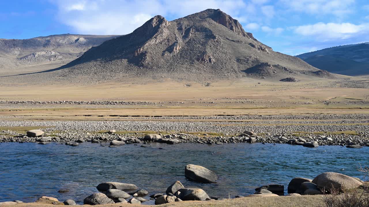 A tranquil autumn scene in an untouched Mongolian valley. The reflection of a rugged mountain shimmers on the clear, pristine water of a slow-moving river. A serene and picturesque view
