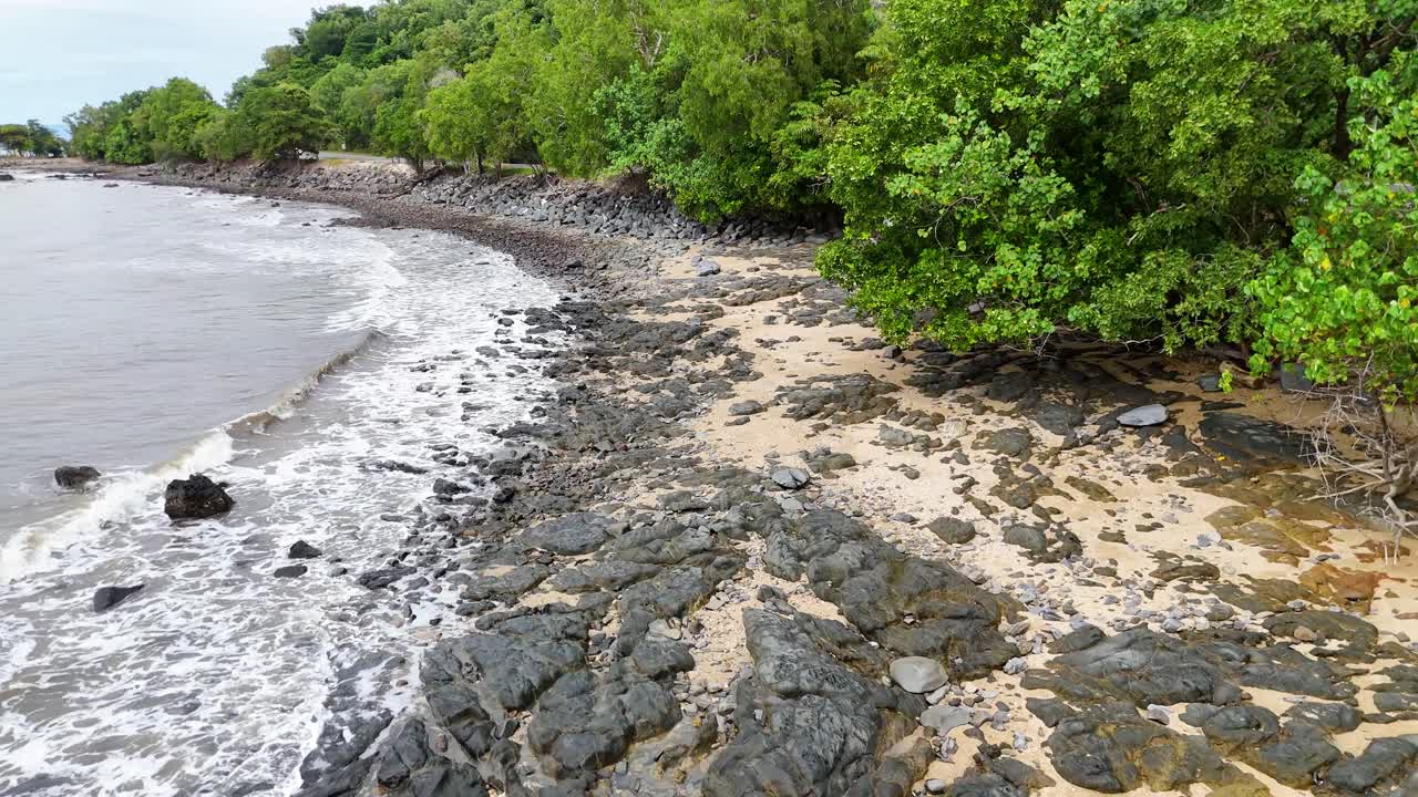 Drone glides above rocky shoreline, ocean waves, and lush green trees under overcast daylight