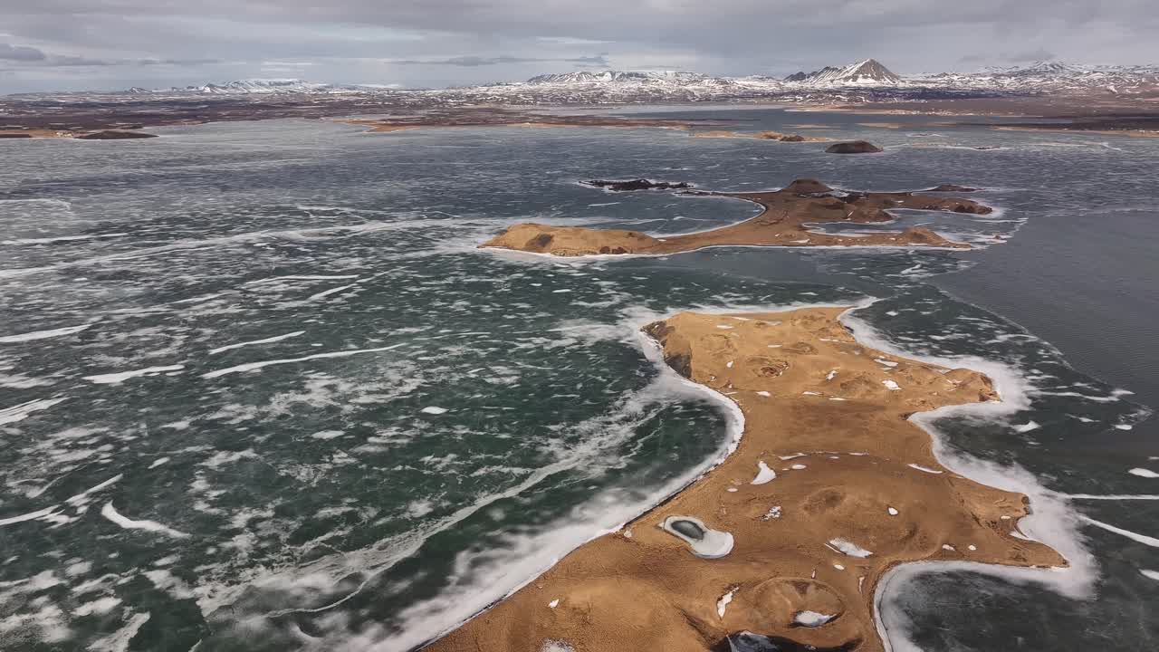 Aerial view of the partially frozen Lake Mývatn, Iceland, showcasing unique volcanic landforms, islands, and distant snowy mountains under a vast sky.