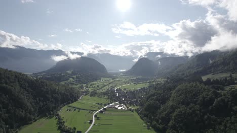 frontal drone video moving over farms in the triglav with hills on the horizon, the sun in front and clear sky with clouds