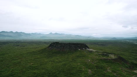 Aerial-volcano-crater-flying-push-in,-scenic-landscape-of-Iceland-Island-during-bright-hazy-day-sunlight-and-blue-sky,-green-field,-volcanic-meadow,-eruption-zone,-old-lava-fields