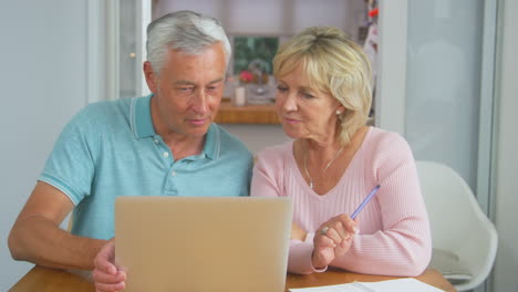 senior retired couple sitting at table at home reviewing personal finances on laptop