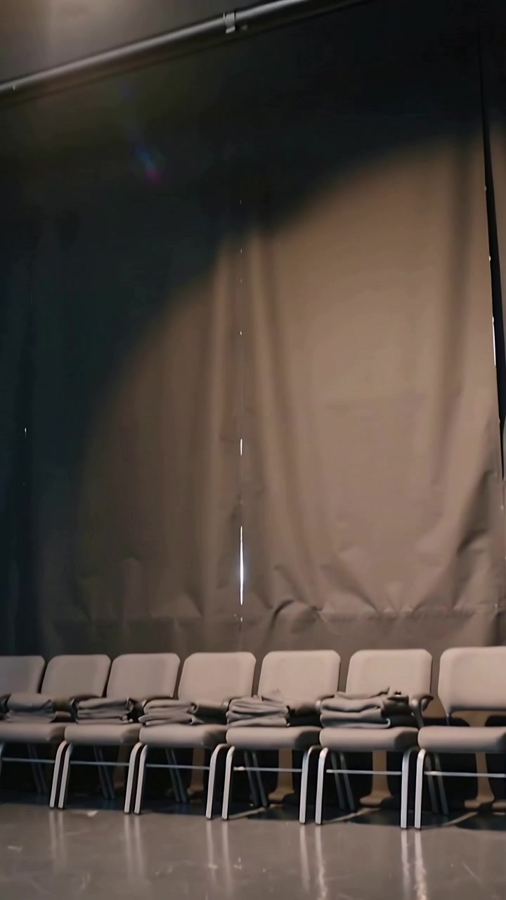 Video verticale: La telecamera si inclina, rivelando un sipario nero, sedie grigie, coperte e un faretto sul palco.