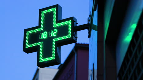 a pharmacy and illuminated international symbol hanging on a corner of a building with a twinkling clock in the middle of a green cross depicting a pharmacy caputred in 4k 60fps