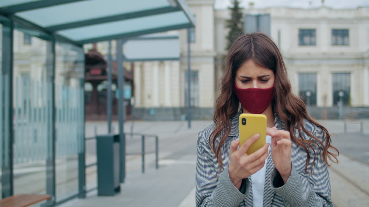 Young Woman in Face Mask Using Smartphone at a Public Transport Stop