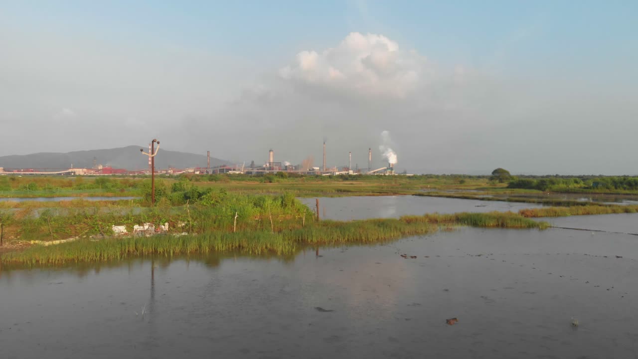 Drone dolly forward drone shot over Waterlogged Indian rice paddy - fish farming fields towards industrial factory sunset