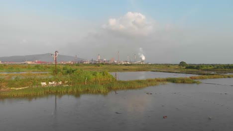 Drone-dolly-forward-drone-shot-over-Waterlogged-Indian-rice-paddy---fish-farming-fields-towards-industrial-factory-sunset