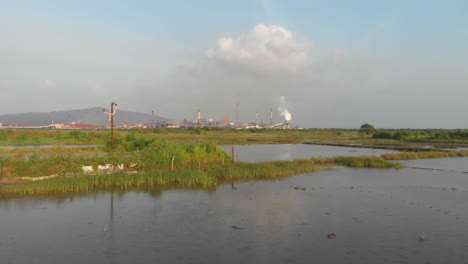 Drone dolly forward drone shot over Waterlogged Indian rice paddy - fish farming fields towards industrial factory sunset