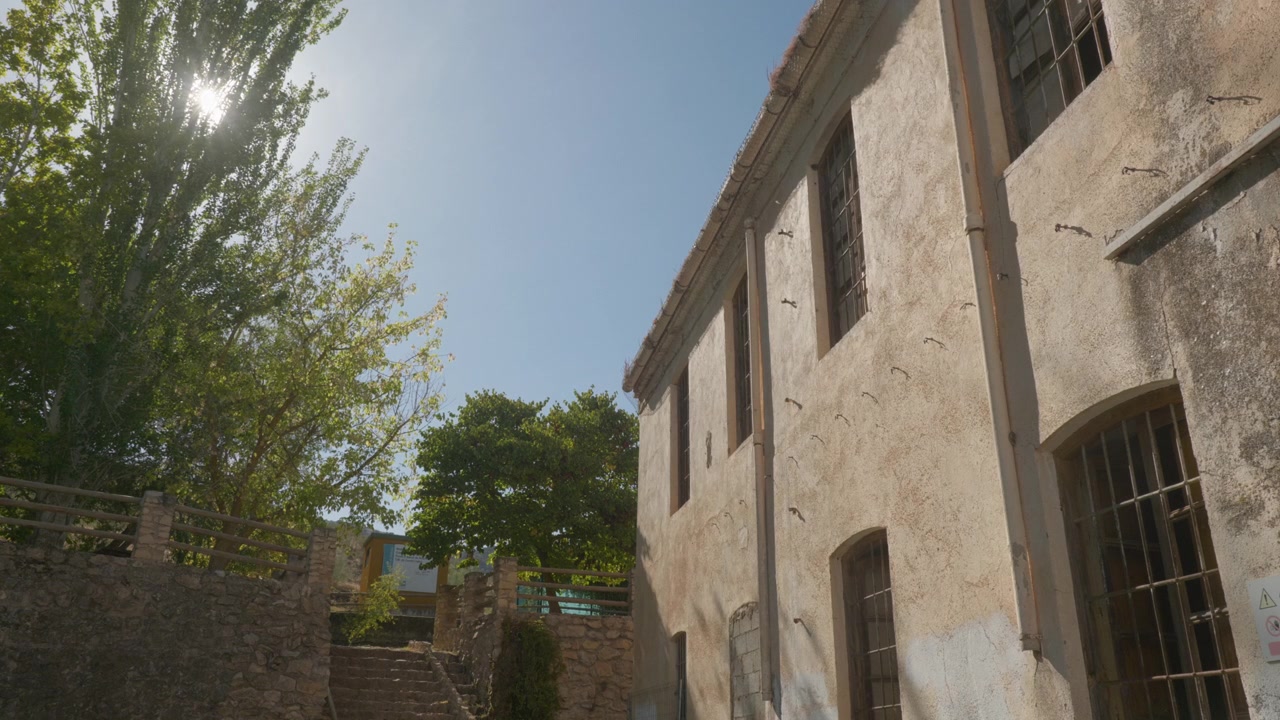 Una vecchia facciata di edificio industriale, logora, con finestre sbarrate, si erge accanto a una scalinata in pietra e alberi verdi sotto un cielo limpido e soleggiato. Ideale per concetti di architettura abbandonata, patrimonio industriale.