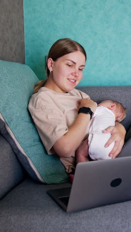 new-made mom with baby in hands sits on sofa. caucasian woman working remote at home combining work and childcare. vertical video