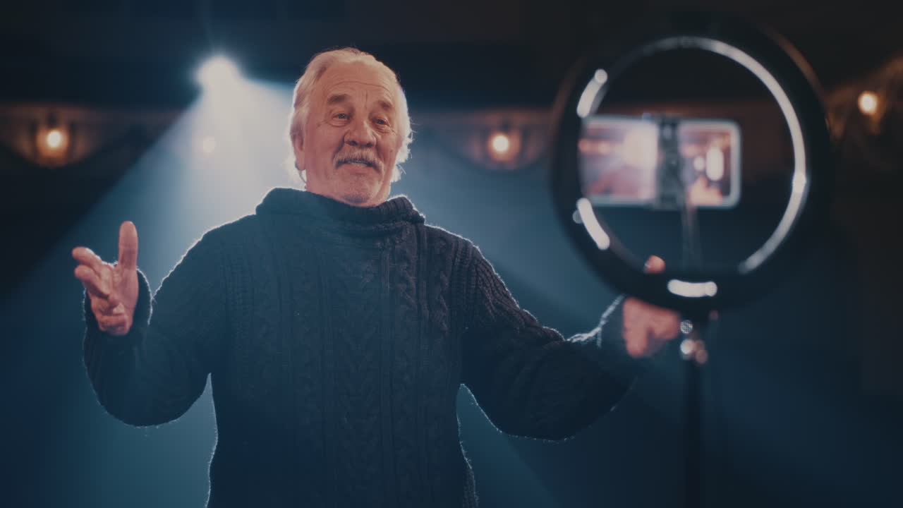 Aged Actor Recording Video on Theater Stage Rack Focus of Elderly Man in Sweater Gesticulating and Speaking to Online Audience while Recording Video in Spotlight in Theater