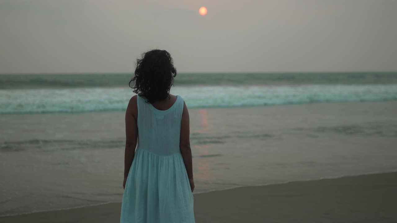 joven con vestido blanco de pie en la playa al atardecer, viendo las olas