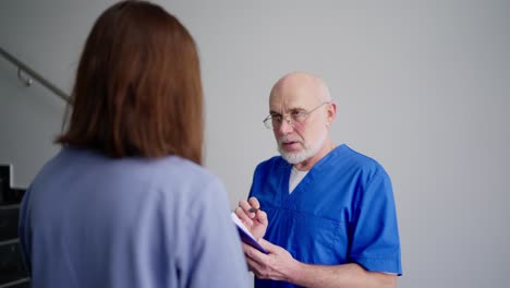 Über-Seiner-Schulter-Erzählt-Ein-Ernster-Arzt-Mit-Brille-Und-Grauem-Bart-In-Blauer-Uniform-Brünetten-Mädchen-Seine-Vorstellungen-über-Symptome-Und-Diagnosen-In-Einer-Modernen-Klinik