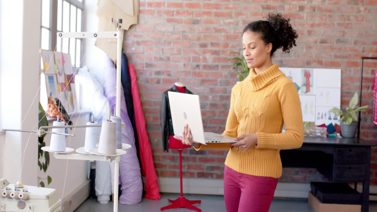 Créatrice de mode biraciale heureuse utilisant un ordinateur portable debout dans un studio, espace pour le texte, ralenti