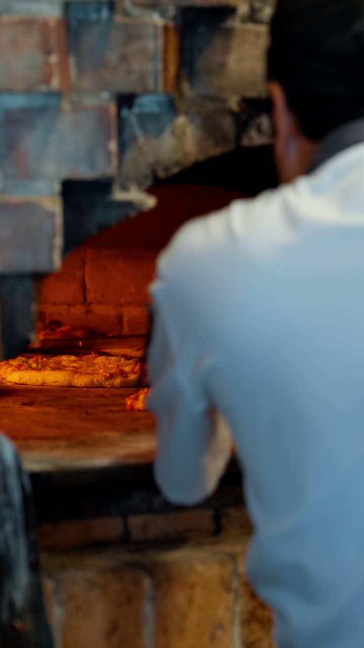 Slow motion vertical close-up of a chef using a peel to take a freshly baked pizza out of a traditional wood-fired brick oven
