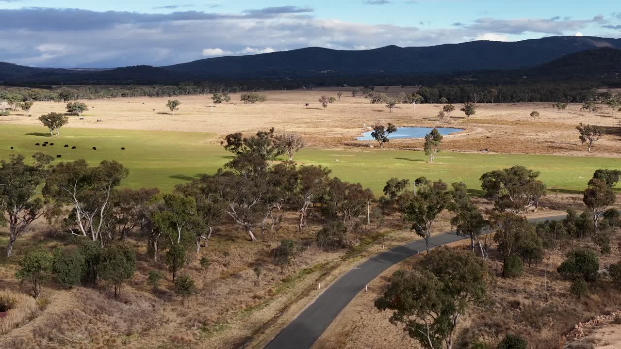 Drone footage glides above a winding rural road, passing trees, ponds, and open fields under a cloudy sky in Stanthorpe, Queensland, Australia