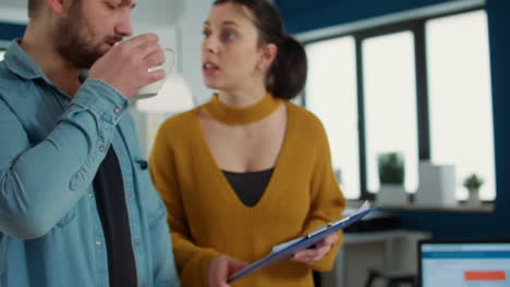 primer plano de la mano del empleado recogiendo una taza de café o té y bebiendo mientras habla con un compañero de trabajo sosteniendo el clipboard