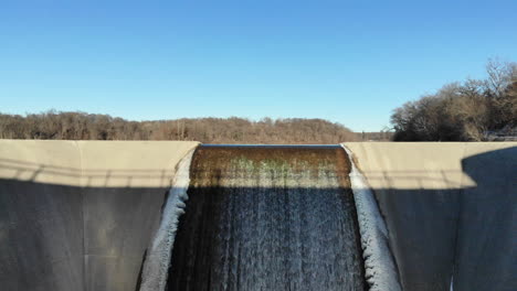 drone flying under a bridge up and over a dam spillway out across a partially frozen lake in winter