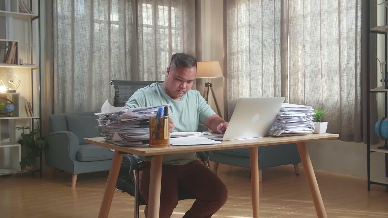 hombre gordo asiático usando una computadora portátil y luego tomando notas mientras trabaja con documentos en la oficina
