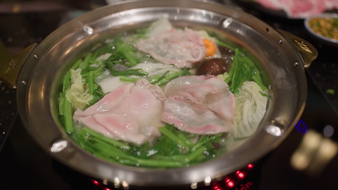 Chopsticks add raw pork slices to boiling hot pot with vegetables under warm, indoor lighting