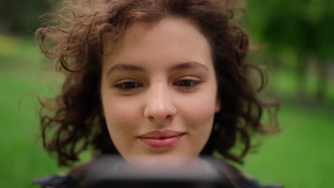 Close-up-a-girl-with-curly-hair-with-a-piercing-on-her-nose-communicates-on-social-networks-while-relaxing-in-the-park