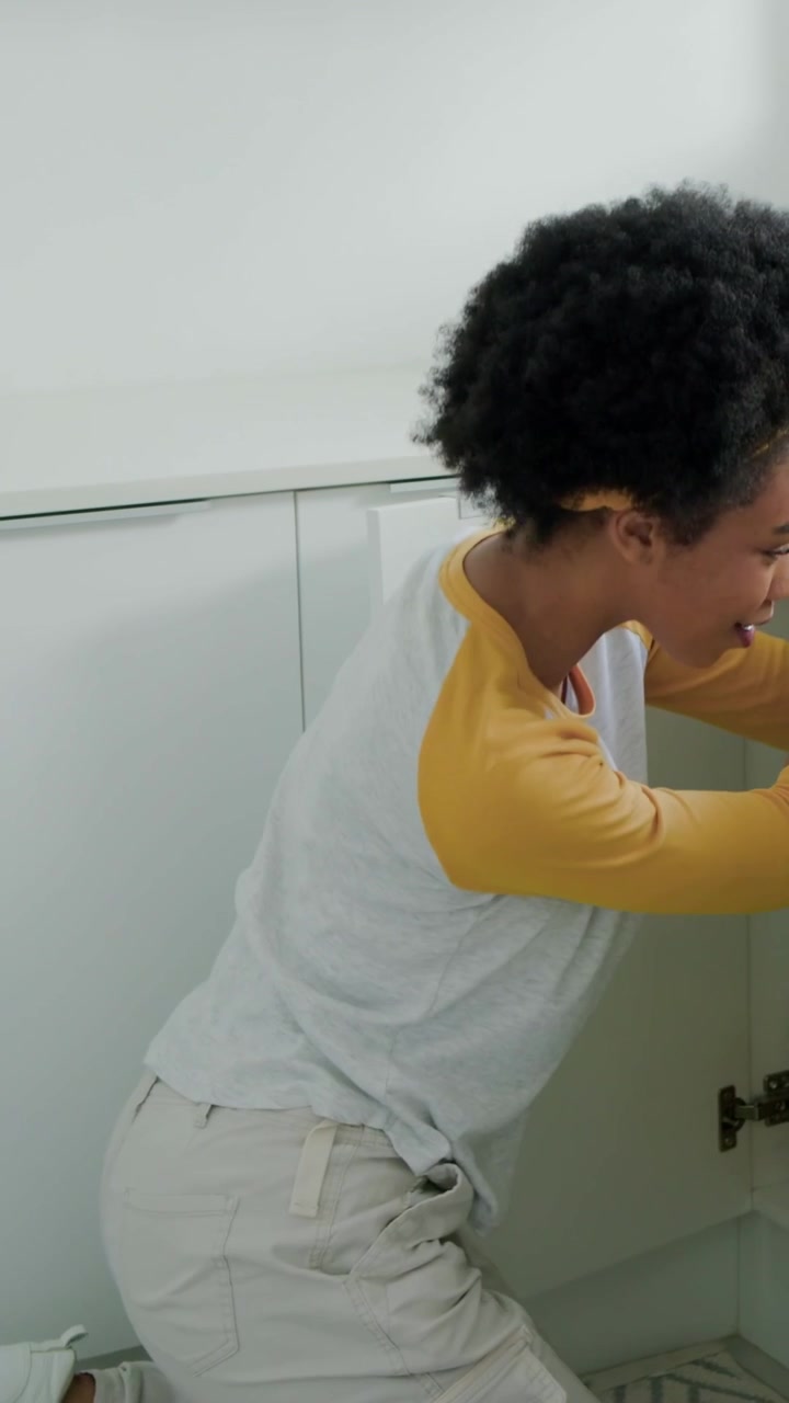 Vertical video: Kneeling woman opening sink cabinet in kitchen checking plumbing with yellow band