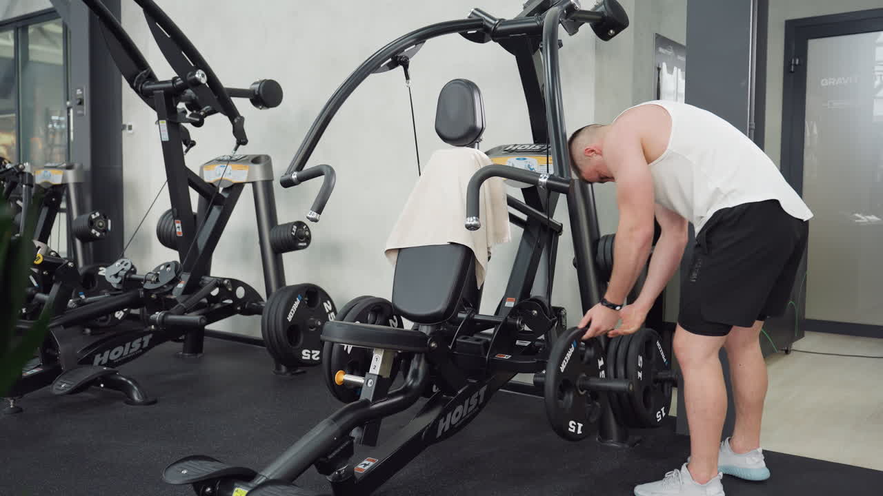 Muscular man removes weight plate from back of weight machine and places it on front for balance while engaging muscles in gym environment under bright lighting with focused strength and intensity