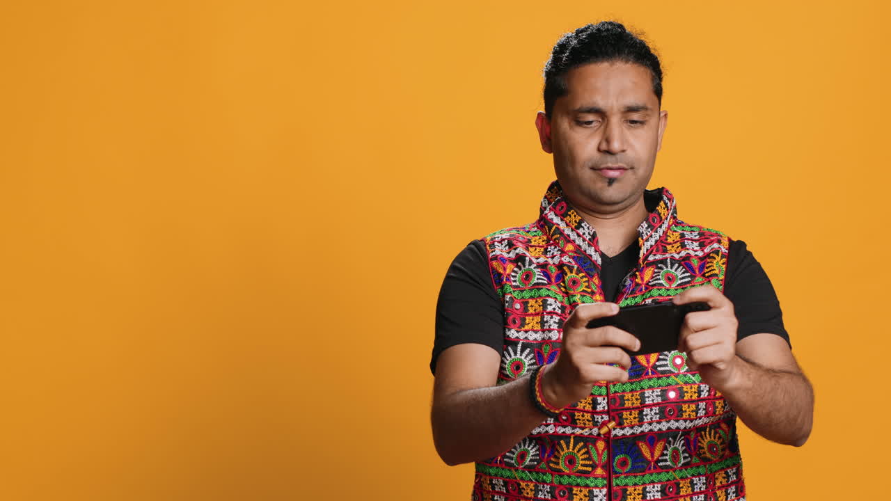 전통 의 옷 을 입은 즐거운 남자 가 드폰 에서 비디오 게임 을 즐기고 있다