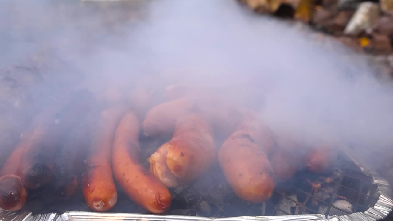 Tampilan close up sosis cokelat keemasan dan hot dog yang dimasak di atas panggangan instan atau barbekyu di luar ruangan selama hari musim gugur dengan asap tebal putih bergerak perlahan dalam gerakan lambat tertiup angin. Latar belakang bokeh