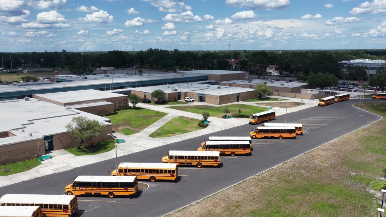 Yellow school buses queue in long rows across paved parking lot outside high school campus in Plant City, Florida, tan brick buildings sit beneath scattered white clouds. Aerial drone rising wide shot