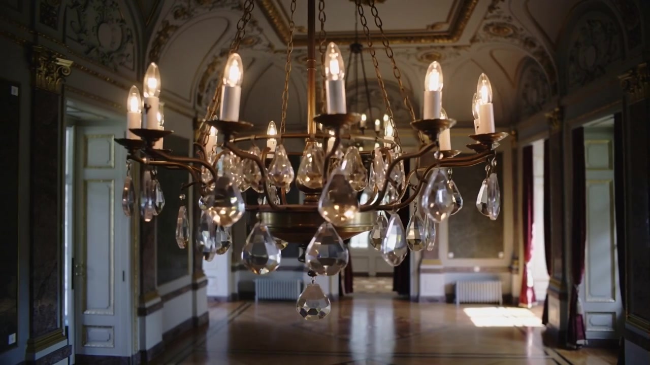 Telecamera in movimento che si avvicina a un lampadario di ottone ornato nel grande salone, rivelando lampadine accese e cristalli.