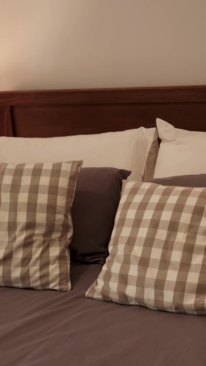 Delle mani posizionano cuscini a quadri su un letto ben fatto in una camera da letto con luce soffusa, creando un'atmosfera calda e accogliente.