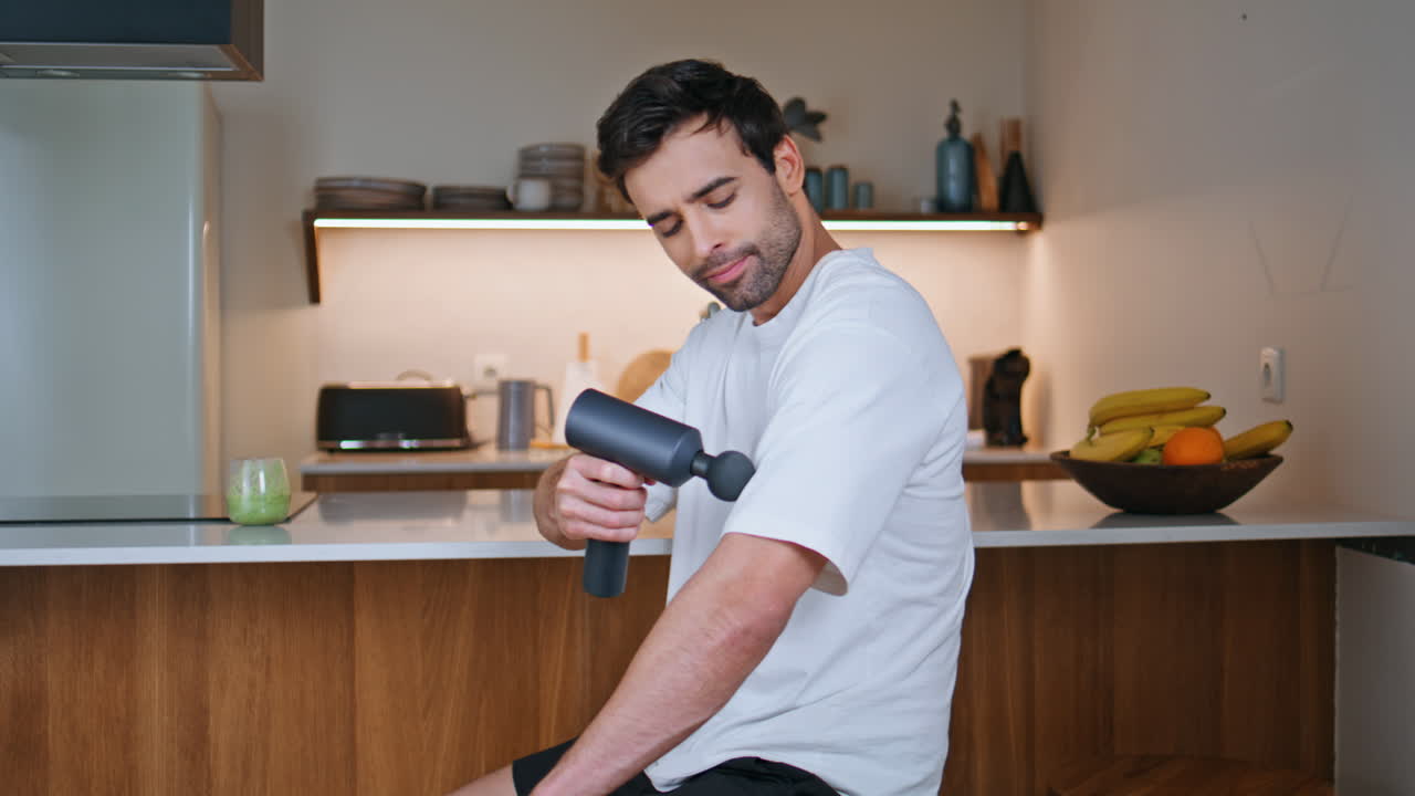Sportsman applying massage gun to shoulder sitting kitchen closeup. o man