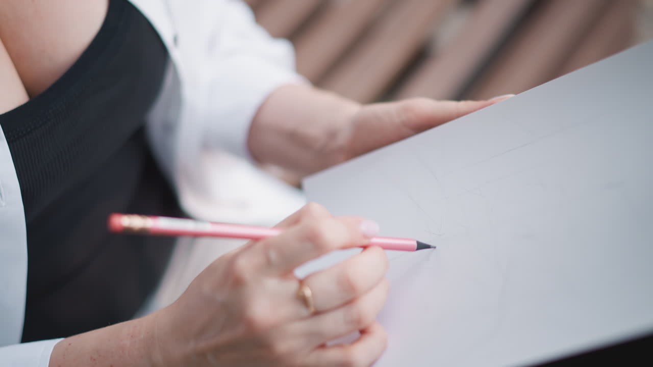 Primo piano delle mani di una donna che disegna su un blocco, anelli d'oro visibili, indossa un top nero e una camicia bianca, sullo sfondo una panchina di legno, tratti di matita delicati, composizione focalizzata, routine personale, marcatura precisa.