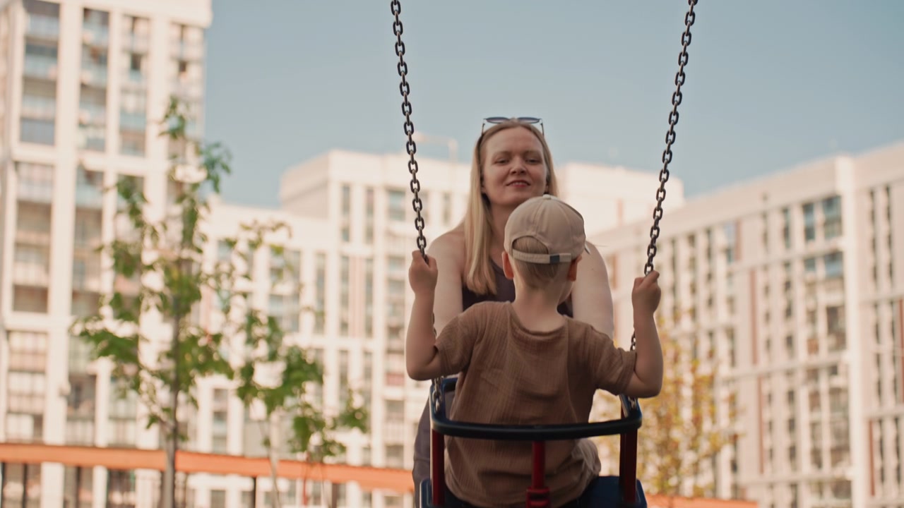 Una donna spinge delicatamente un bambino sull'altalena in un parco giochi all'aperto. La scena cattura l'interazione tra loro con alti edifici e vegetazione visibili sullo sfondo. Una luce naturale soffusa illumina