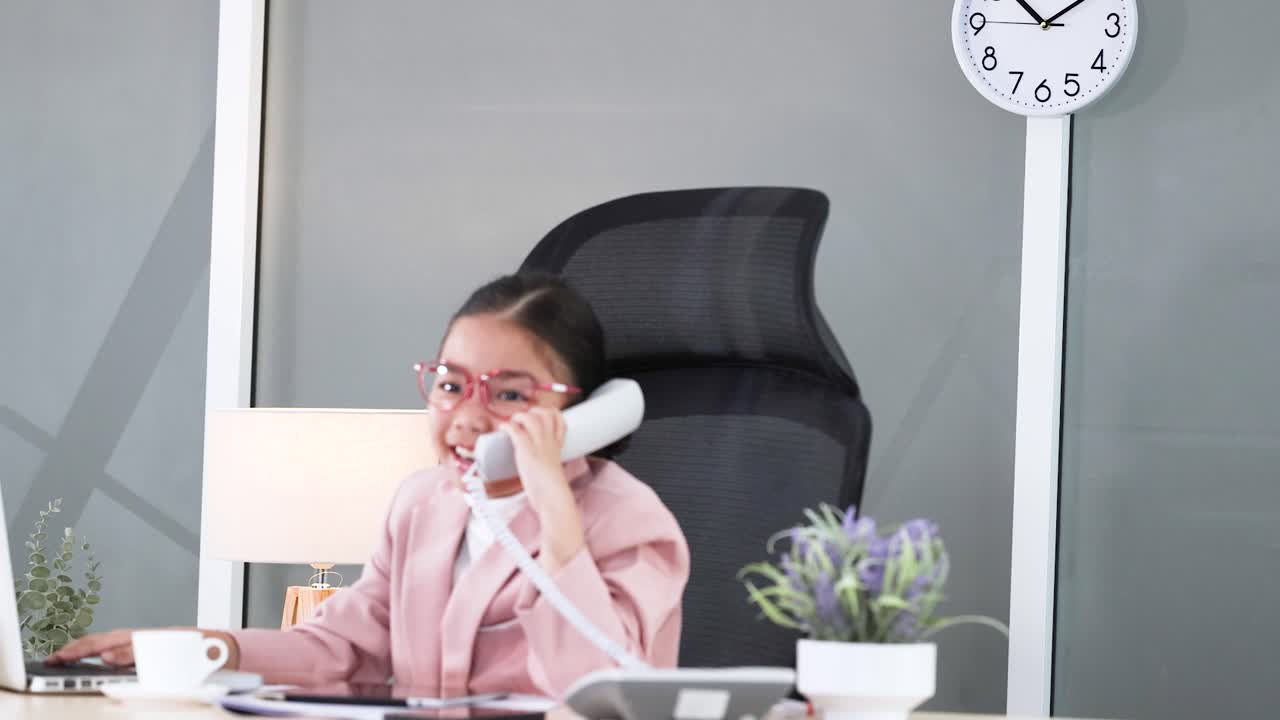 Una giovane ragazza in tailleur rosa parla al telefono a una scrivania d'ufficio.
