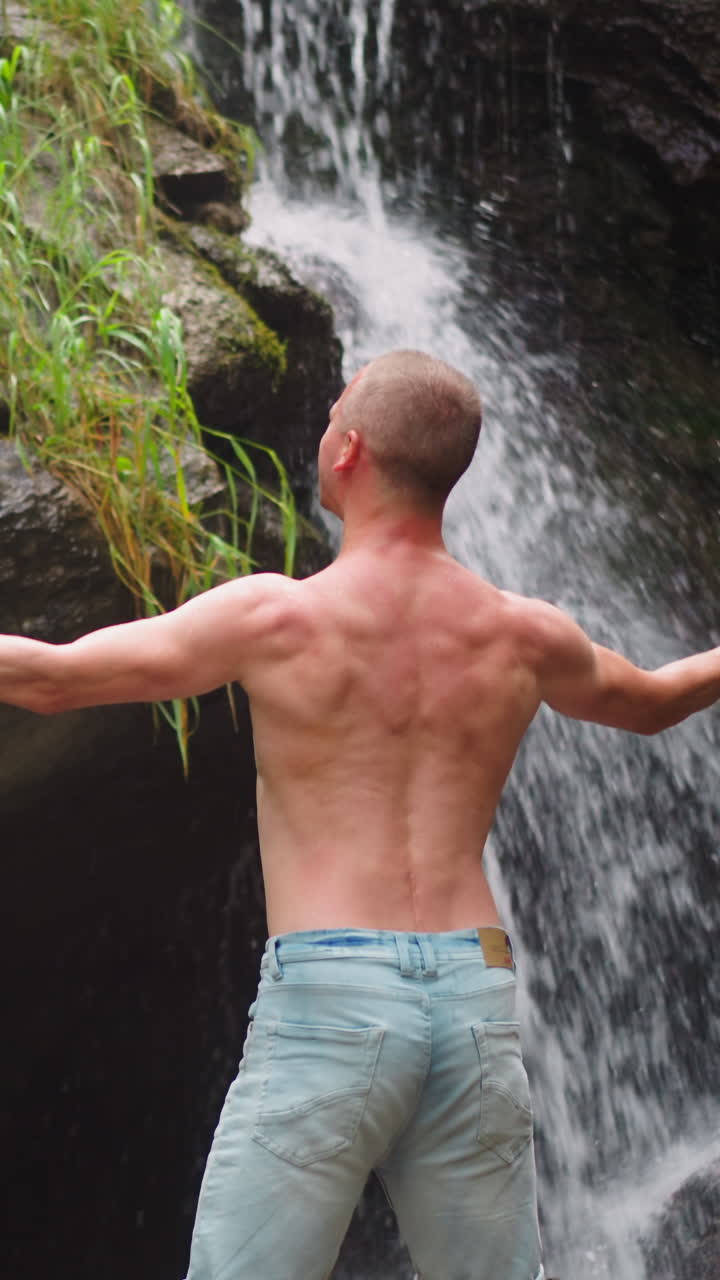 hombre sin camisa levanta las manos feliz de vincularse con la naturaleza virgen cerca de cascadas y grandes rocas en las tierras altas vista trasera en cámara lenta