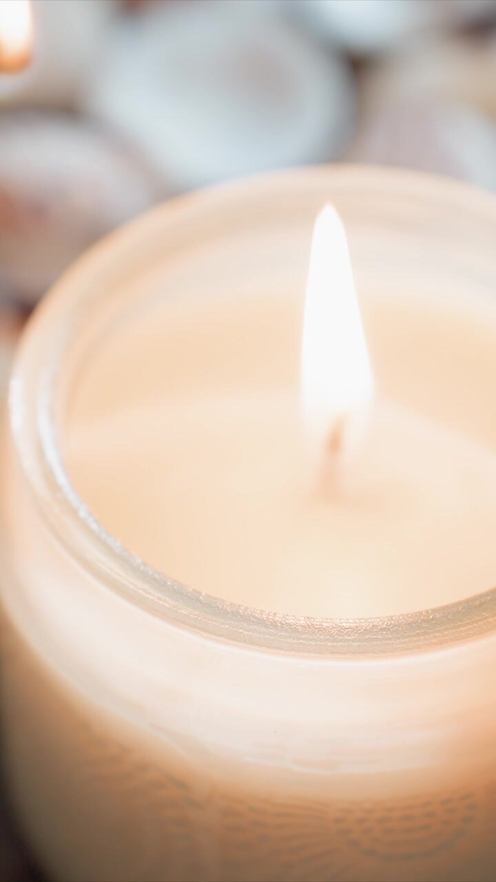 Nærbillede af en hånd, der holder og tænder et duftfrit lys med blød hvid voks i et klart glas. Flammen antændes gradvist og kaster varmt lys på de glatte teksturdetaljer omkring vægeområdet. Baggrund.