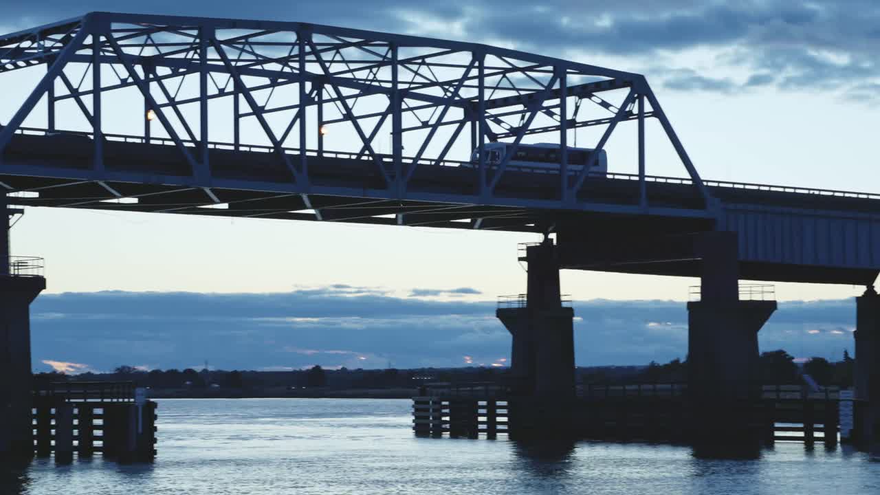 A semi-truck drives across a bridge over water at twilight with a dramatic blue sky and scattered clouds