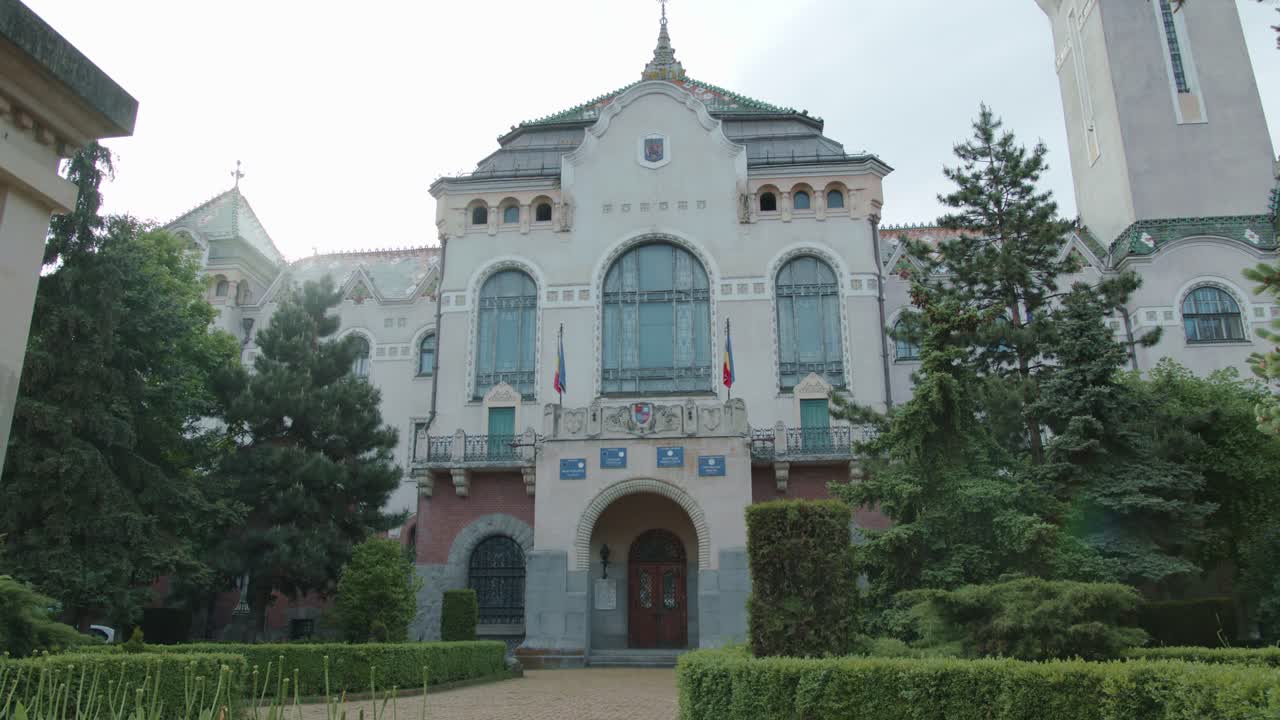 A wide, static establishing shot of the ornate Art Nouveau facade of the Palace of Culture in Targu Mures. The historic building is seen from its garden on a cloudy day