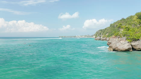 Low-aerial-view-of-surfers-in-tropical-blue-ocean-by-steep-cliffs-with-luxury-villas-in-Bali,-Indonesia