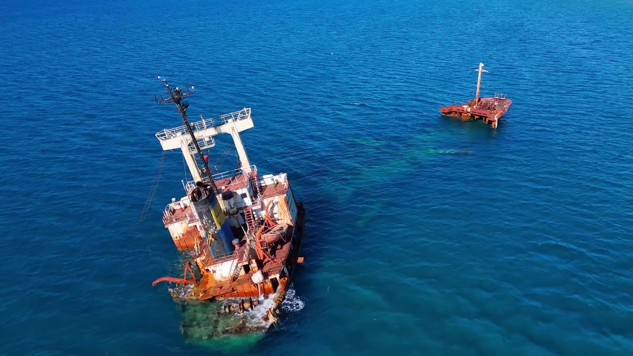 The drone advances toward the tilted main hull of the Manassa shipwreck off Crete, revealing corroded decks, a leaning mast and the detached bow across clear blue water.