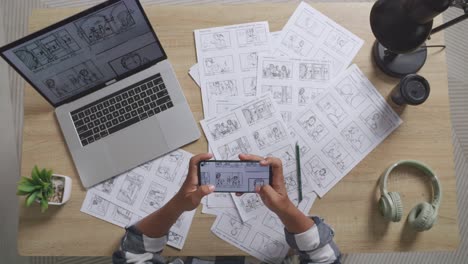 vista superior zoom out toma de artista masculino asiático mirando el concepto de dibujo en el teléfono inteligente mientras dibuja el guión gráfico en la mesa con una computadora portátil y auriculares en el estudio