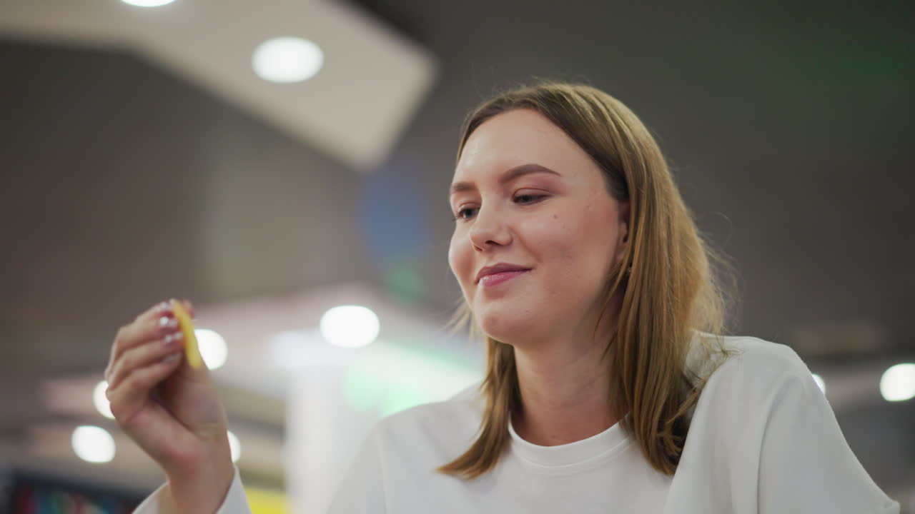 primer plano de una mujer comiendo papas fritas con una sonrisa cálida, mirada enfocada, luces de colores parpadeando en el fondo, disfrutando de un bocadillo en un ambiente de comedor vibrante y casual con una atmósfera alegre