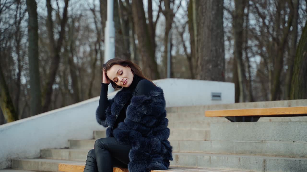 Luxury girl sitting on bench. Attractive young woman in fur coat enjoys fresh air in the park and smiles. Positive brunette outdoors.